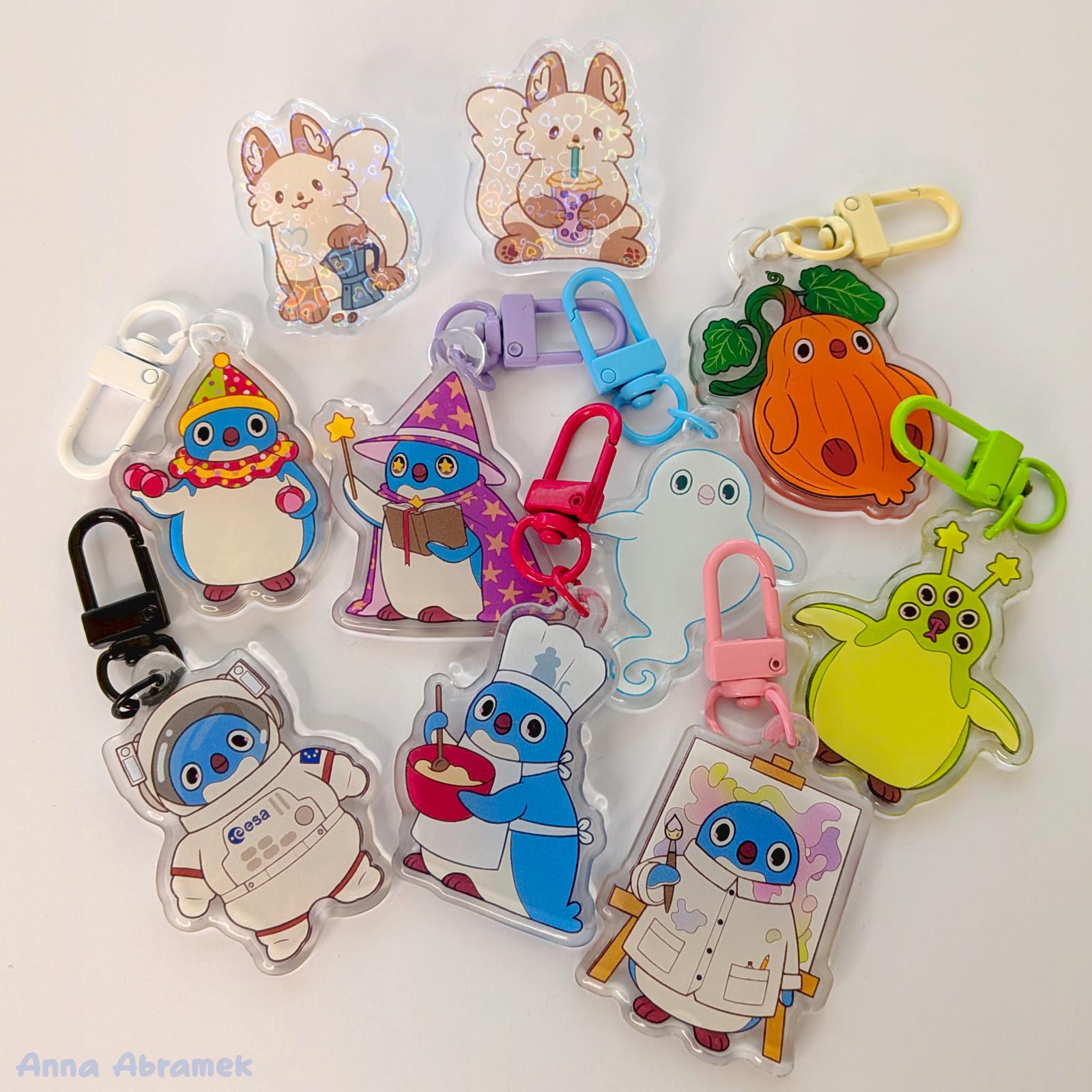 A collection of acrylic keychains with drawings of penguins, and acrylic pins featuring arctic foxes, laid out on a white background.